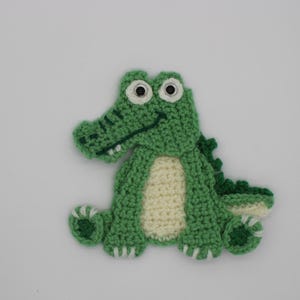 May include: A crocheted alligator applique in green and cream. The alligator has white eyes, a smiling mouth, and a textured body. This whimsical applique is ideal for adding a playful touch to clothing or accessories.
