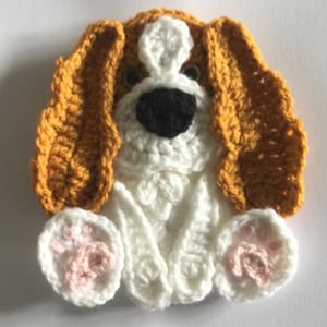 May include: A crocheted dog applique, featuring a white body, black nose, and brown ears. The paws are white with pink paw pads. The dog is in a sitting position. The eyes are small and black.