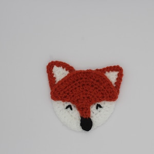 May include: A crocheted fox face with a white face, orange head, and black nose and eyes. The fox has pointed ears with white inner ears. The craft is made with yarn and is likely a decorative item or applique.