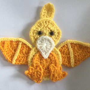 May include: A handmade crochet pterodactyl in shades of yellow and orange. The wings, body, and head are crafted with textured yarn, featuring a white beak and black safety eyes. This whimsical creation is perfect for a nursery or as a unique gift.