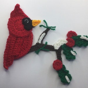 May include: A handmade crochet cardinal bird with a black eye, yellow beak, and vibrant red feathers. The bird is perched on a brown branch with green leaves, white accents, and red berries. This decorative piece is perfect for holiday or nature-themed decor.