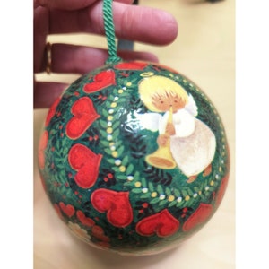 May include: A green Christmas ornament with a painted image of an angel playing a trumpet. The ornament is decorated with red hearts and green leaves.
