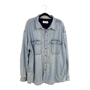 May include: A light blue, acid-washed denim long-sleeve shirt featuring a dark navy blue inner collar lining, a button-down front with metal buttons, and two chest pockets with buttoned flaps. The shirt has a collar and an Orvis brand label.