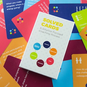 Op de afbeelding: Een set kleurrijke kaarten met een witte kaart in het midden waarop "SOLVED CARDS" en "A Solution Focused Coaching Process" staat. De witte kaart bevat een diagram van een cirkel met zes secties die zijn gelabeld met "Exception", "Evaluation", "Level", "Intervention", "Measurement & Direction" en "Execution".
