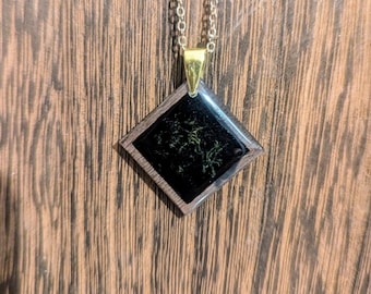 May include: A black diamond-shaped pendant with a gold chain. The pendant has a black resin inlay with a green pattern.