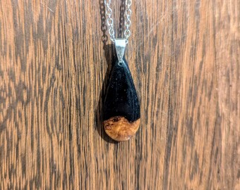 May include: A teardrop-shaped pendant necklace with a black and brown wood inlay. The pendant is attached to a silver chain.