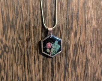 May include: A gold chain necklace with a hexagonal pendant featuring a pressed flower and greenery encased in clear resin.