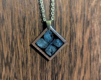 May include: A gold chain necklace with a square pendant featuring a pressed flower encased in resin. The pendant is made of dark wood with a black border.