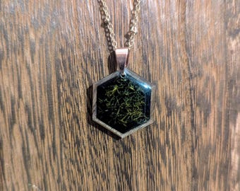 May include: A gold chain necklace with a hexagonal pendant. The pendant is filled with black resin and has a gold-colored wire design inside.
