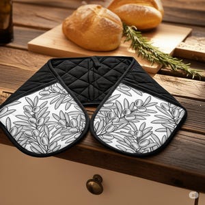 Puede incluir: Manopla de horno acolchada en blanco y negro con diseño de rama de olivo. La manopla tiene un borde negro y un lazo para colgar. El fondo incluye pan y romero.