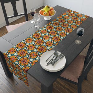 May include: A colourful table runner with a geometric pattern in shades of orange, yellow, blue, and green. The runner is laid on a dark wood table with a white plate and cutlery.