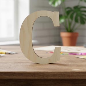 May include: A wooden letter "C" stands on a wooden surface. The letter is light-colored and has a simple, clean design. The background includes a window, a potted plant, and various stationery items.