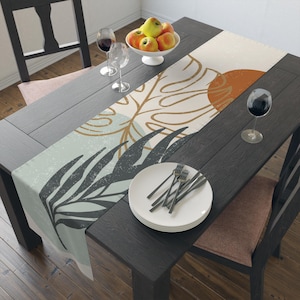 May include: A decorative table runner with a botanical design. The runner features a light blue, beige, and orange color palette with leaf and sun illustrations. A white plate with forks and a bowl of apples sit on a dark wooden table.