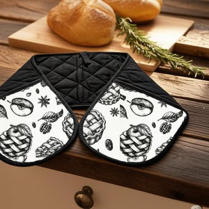 Puede incluir: Manopla de horno en blanco y negro con una parte superior negra acolchada y una parte inferior estampada en blanco. El estampado presenta ilustraciones detalladas de manzanas, tartas de manzana, hojas y nueces. La manopla está diseñada para proteger las manos del calor.