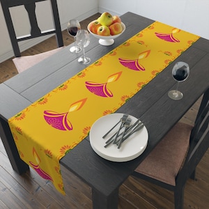 May include: A yellow table runner with a repeating pattern of pink and orange diya lamps. The runner is on a dark wood table with white plates and silverware.