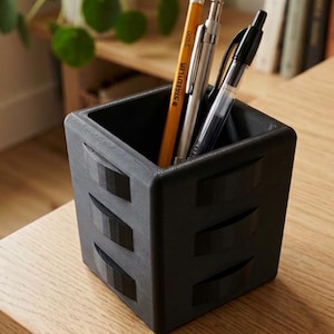 Puede incluir: Un organizador de escritorio negro, en forma de cubo, con tres cortes horizontales decorativos en cada lado. El organizador contiene un lápiz amarillo, un portaminas plateado y un bolígrafo negro. Se encuentra sobre una superficie de madera clara.