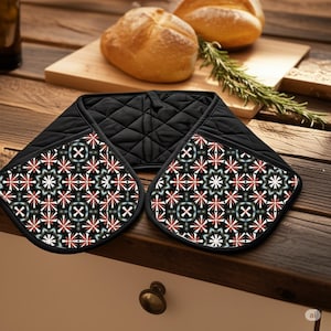 Puede incluir: Manopla de horno acolchada negra con un estampado floral decorativo en rojo, blanco y verde azulado. La manopla está diseñada para proteger las manos del calor al manipular utensilios de cocina calientes. Sobre una superficie de madera con pan y hierbas.