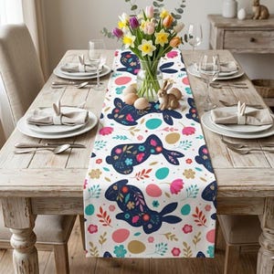 May include: A white Easter table runner with a repeating pattern of navy blue floral bunnies, colorful eggs, and floral accents. The runner is set on a wooden table with place settings, a vase of flowers, and a decorative bunny figurine.