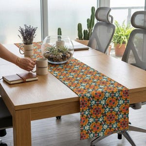 May include: A wooden table with a colorful floral patterned table runner. The runner features a repeating design in shades of orange, teal, and white. A person is holding a mug with text. A glass bowl with plants and rocks sits on the table.