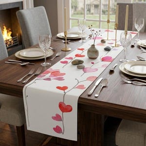 Puede incluir: Una mesa de comedor preparada para una comida, con un camino de mesa blanco con un diseño de corazón en acuarela en tonos rojos y rosas. La mesa está puesta con platos, cubiertos y copas de vino, con velas y un pequeño jarrón.