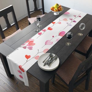 May include: White table runner with a watercolor design of pink and red hearts on branches. The runner is on a dark wood table with two chairs and a bowl of fruit.