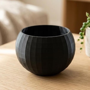 Puede incluir: Una maceta negra, esférica, con detalles de costillas verticales. La maceta tiene una abertura ancha y una superficie texturizada. Está sobre una superficie de madera clara.