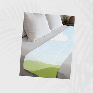 May include: A white quilted bed runner with a light blue and green gradient design. The runner is placed on a gray bed with white pillows. The design features a heart shape.