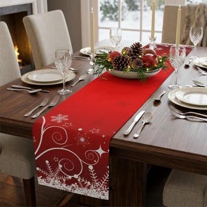 Puede incluir: Una mesa navideña festiva con un camino de mesa rojo con diseños de copos de nieve y remolinos blancos. La mesa está puesta con platos, cubiertos y copas de vino. Un centro de mesa de piñas, manzanas y vegetación se encuentra en un cuenco.