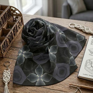 May include: A dark gray scarf with a floral pattern, displayed on a wooden surface. The scarf features a repeating design of stylized flowers and geometric shapes in shades of gray and black. A wooden organizer and a macrame keychain are also visible.