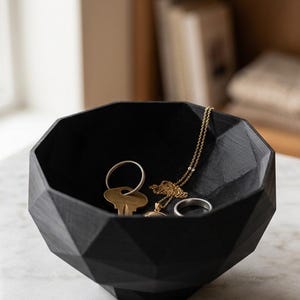 Puede incluir: Un cuenco negro de forma geométrica que contiene llaves, un collar de oro y un anillo de plata. El cuenco tiene un diseño facetado y está sobre una superficie de mármol. La bandeja para joyas es una solución de almacenamiento decorativa.