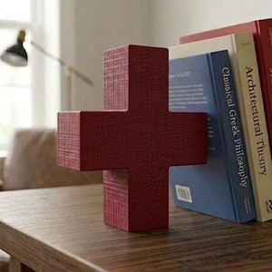 May include: A dark red, cross-shaped bookend with a textured, fabric-like surface finish. It stands upright next to two hardcover books, one with a blue spine titled Classical Greek Philosophy and one with a cream spine titled Architectural Theory.