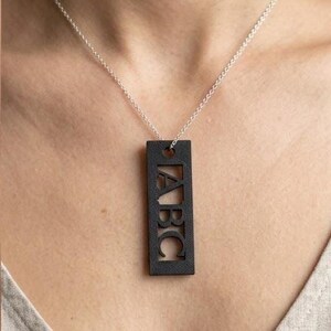 May include: A black rectangular pendant necklace with the letters "ABC" cut out. The pendant hangs from a delicate silver chain. The necklace is worn around the neck.