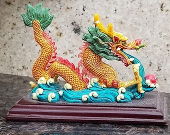 May include: A colorful dragon figurine with green, yellow, and red accents. The dragon is depicted with its mouth open, holding a red orb in its claws. The figurine is set on a wooden base.