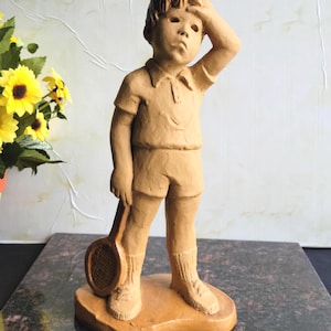 May include: A tan-colored statue of a young person in tennis attire, holding a racket and touching their forehead. The figure wears a collared shirt, shorts, and socks. The statue is on a base.