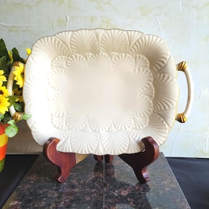 May include: Cream-colored rectangular serving platter with a scalloped edge and gold trim. The platter has a raised, fan-like pattern around the rim and handles. It is displayed on a dark wooden stand, suggesting a decorative or formal use.