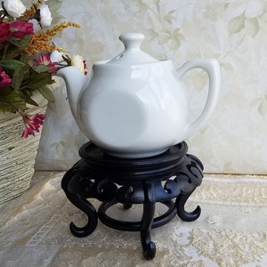 May include: A white ceramic teapot with a lid sits on a black wooden stand with intricate carvings. The teapot has a rounded body and a spout on the side. The stand has three legs and a circular base.