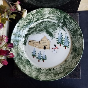 May include: A white ceramic plate with a green rim features a winter scene with a log cabin, pine trees, and a snowman. The plate has a distressed, vintage look.