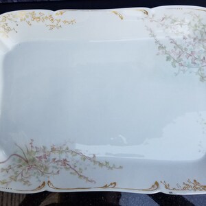 Puede incluir: Una bandeja de servir de porcelana blanca con un borde dorado y un dise&ntilde;o floral. La bandeja tiene dos asas con un dise&ntilde;o de rollo dorado.