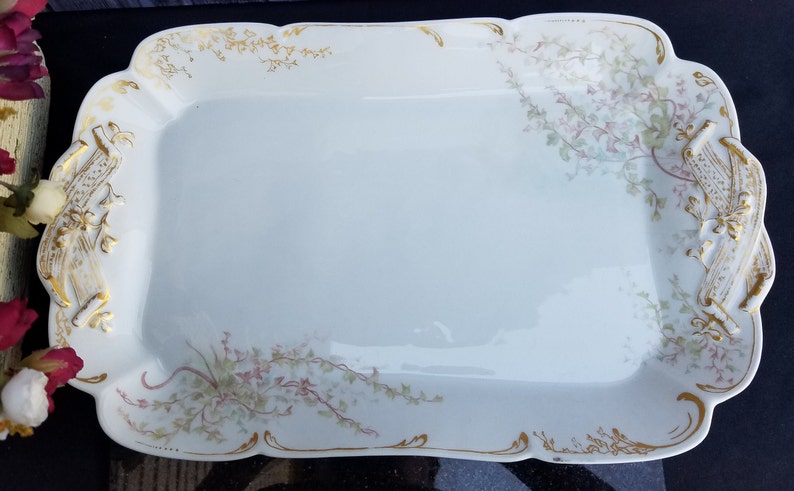 Puede incluir: Una bandeja de servir de porcelana blanca con un borde dorado y un dise&ntilde;o floral. La bandeja tiene dos asas con un dise&ntilde;o de rollo dorado.