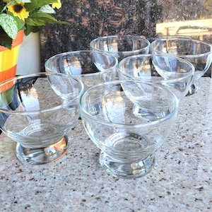 May include: Six clear glass dessert bowls with rounded sides and pedestal bases are arranged on a speckled surface. The bowls are empty and appear to be new. The background includes a colorful flower pot and a dark, textured wall.