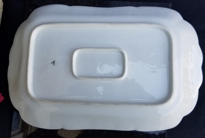 Puede incluir: Plato de servir de porcelana blanca con una secci&oacute;n central elevada y bordes festoneados. El plato tiene una marca del fabricante en la parte inferior que dice "H.F.C.H."