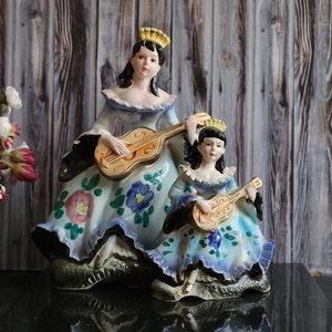 Puede incluir: Dos figuritas de porcelana de mujeres tocando guitarras. La mujer de la izquierda lleva un vestido azul y blanco con motivos florales y una corona. La mujer de la derecha lleva un vestido azul y blanco con motivos florales y una corona. Ambas mujeres sostienen guitarras.