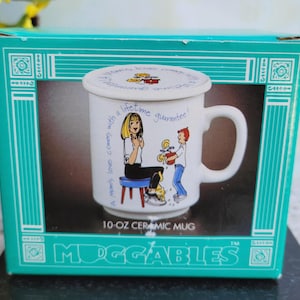 May include: A white ceramic mug with a lid, featuring a cartoon illustration of a woman and a boy. The mug is labeled "10-OZ CERAMIC MUG" and has the text "A Mom's love comes with a lifetime guarantee!" The mug is in a teal box.