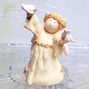 Puede incluir: Figura de ángel color crema sosteniendo dos palomas blancas. El ángel tiene cabello rubio y una cara sonriente. El vestido está adornado con pequeñas rosas rosas. La figura está sobre una superficie de vidrio transparente.