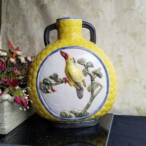 May include: A yellow ceramic vase with a blue and white design featuring a bird on a branch. The vase has two handles and a round body.