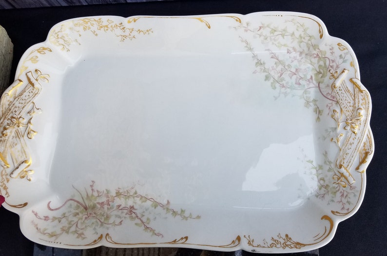 Puede incluir: Una fuente de servir de porcelana blanca con ribete dorado y dise&ntilde;o floral. La fuente tiene dos asas a los lados.