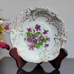 May include: A white porcelain trinket dish with a gold rim and a floral design featuring purple violets. The dish is on a wooden stand.