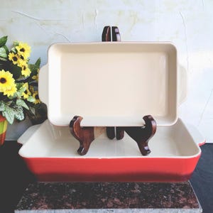 May include: Two rectangular baking dishes. The top dish is cream-colored, resting on a wooden stand. The bottom dish is red with a cream interior. Yellow sunflowers in a colorful pot are in the background.