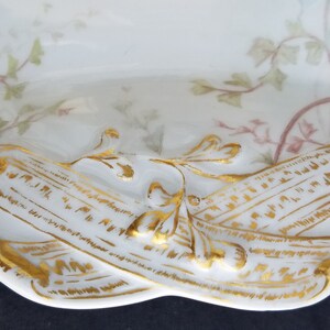 Puede incluir: Un plato de servir de porcelana blanca con un borde dorado y un dise&ntilde;o floral. El plato presenta dos rollos con letras doradas.