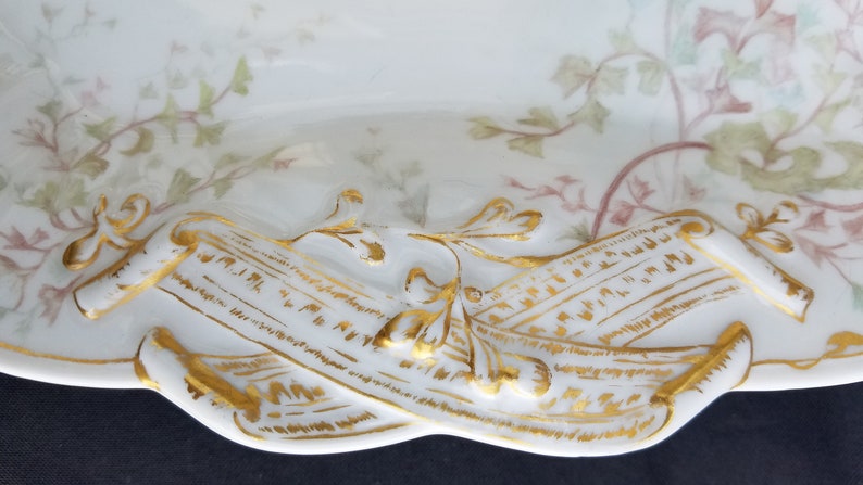 Puede incluir: Un plato de servir de porcelana blanca con un borde dorado y un dise&ntilde;o floral. El plato presenta dos rollos con letras doradas.